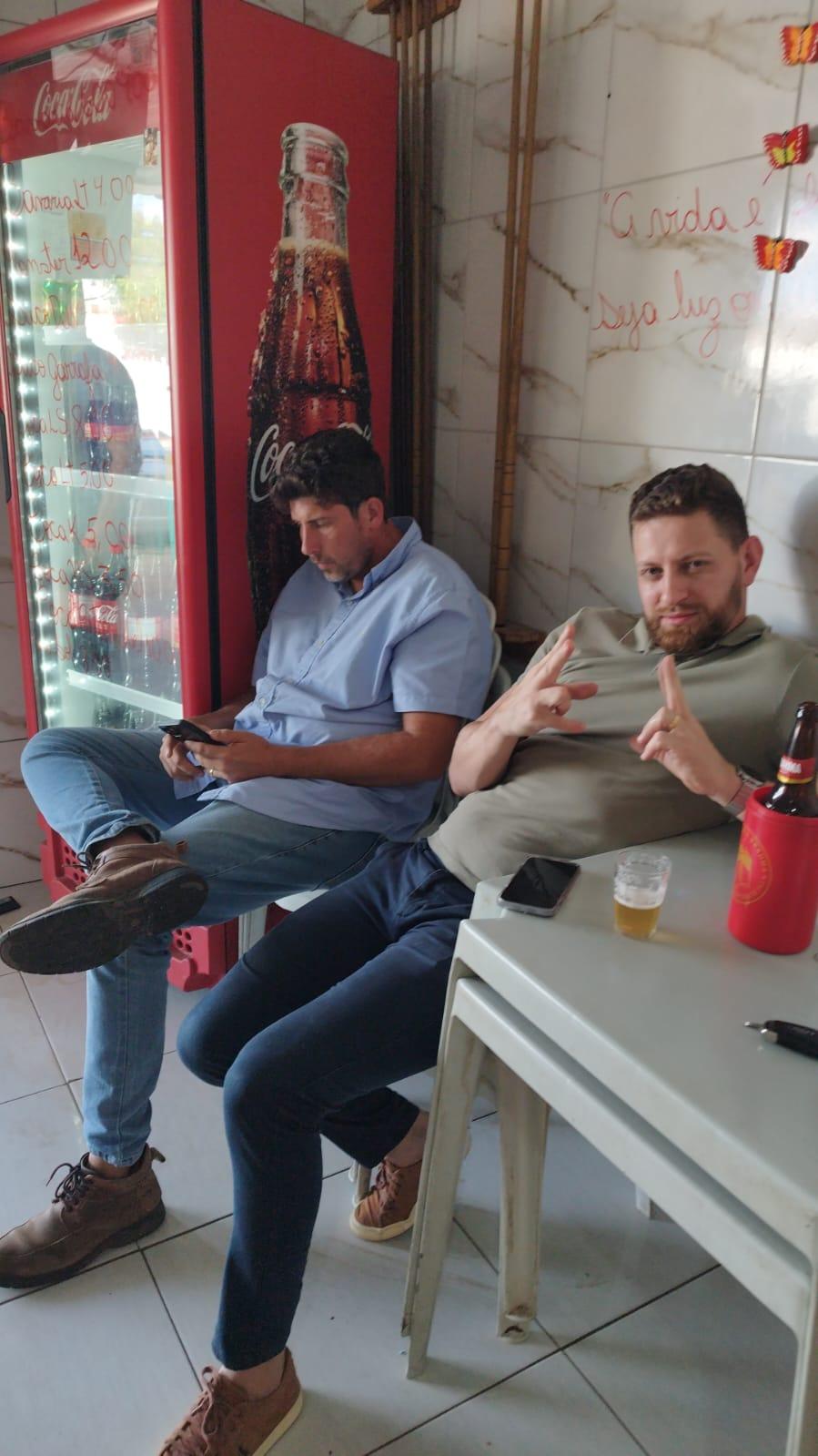Dois amigos no boteco, um mexendo no celular e outro fazendo sinal para a câmera